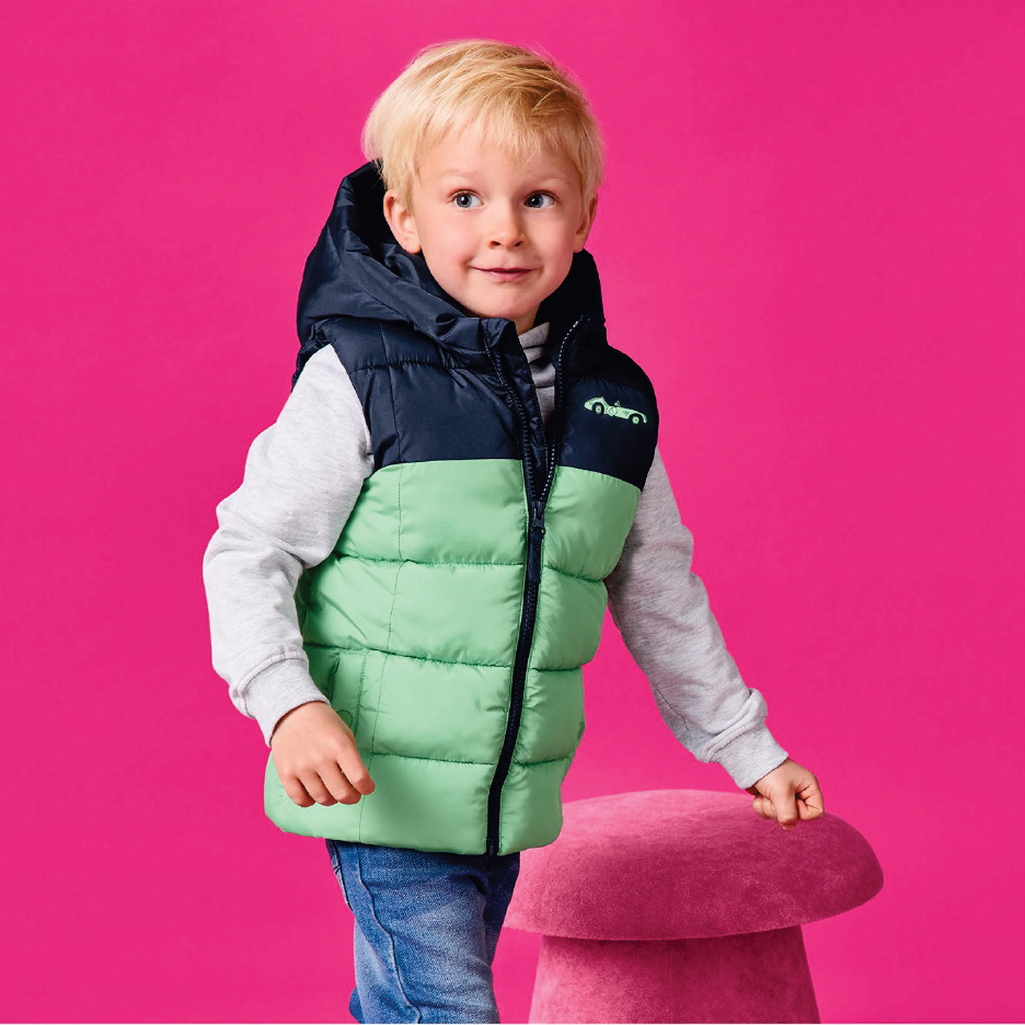 Момче в двуцветен елек с качулка и дълъг ръкав, дънков панталон, докосва розова табуретка.