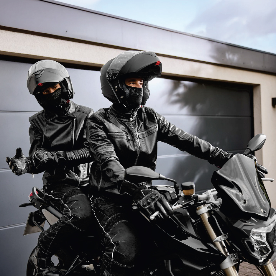 Двама мотористи в черни кожени якета и каски на мотор пред гараж.