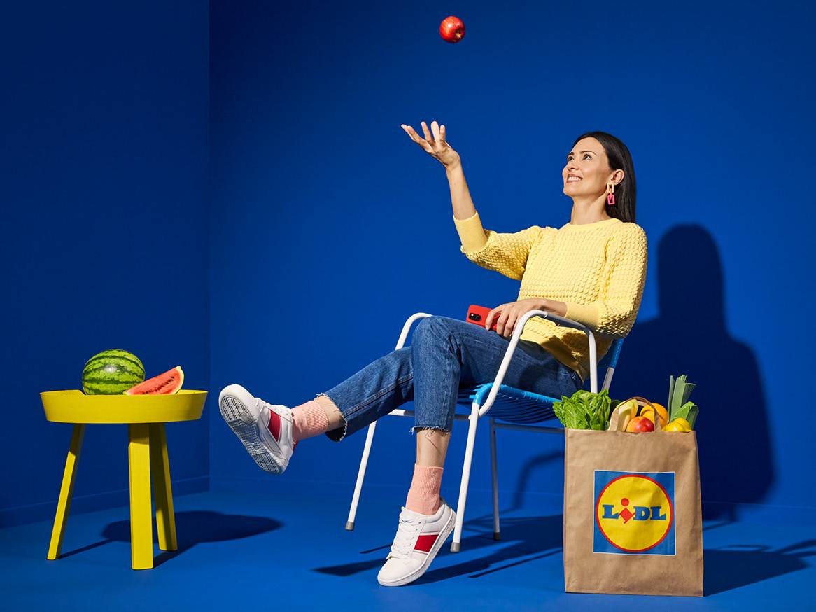 Жена хвърля ябълка във въздуха, седнала на стол с чанта Lidl, пълна с пресни продукти.