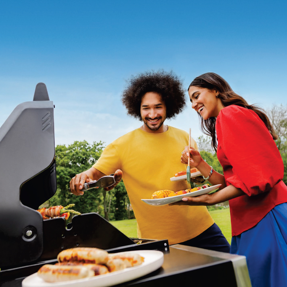 Мъж и жена се усмихват, докато пекат на скара наденички, царевица и шишчета на открито.