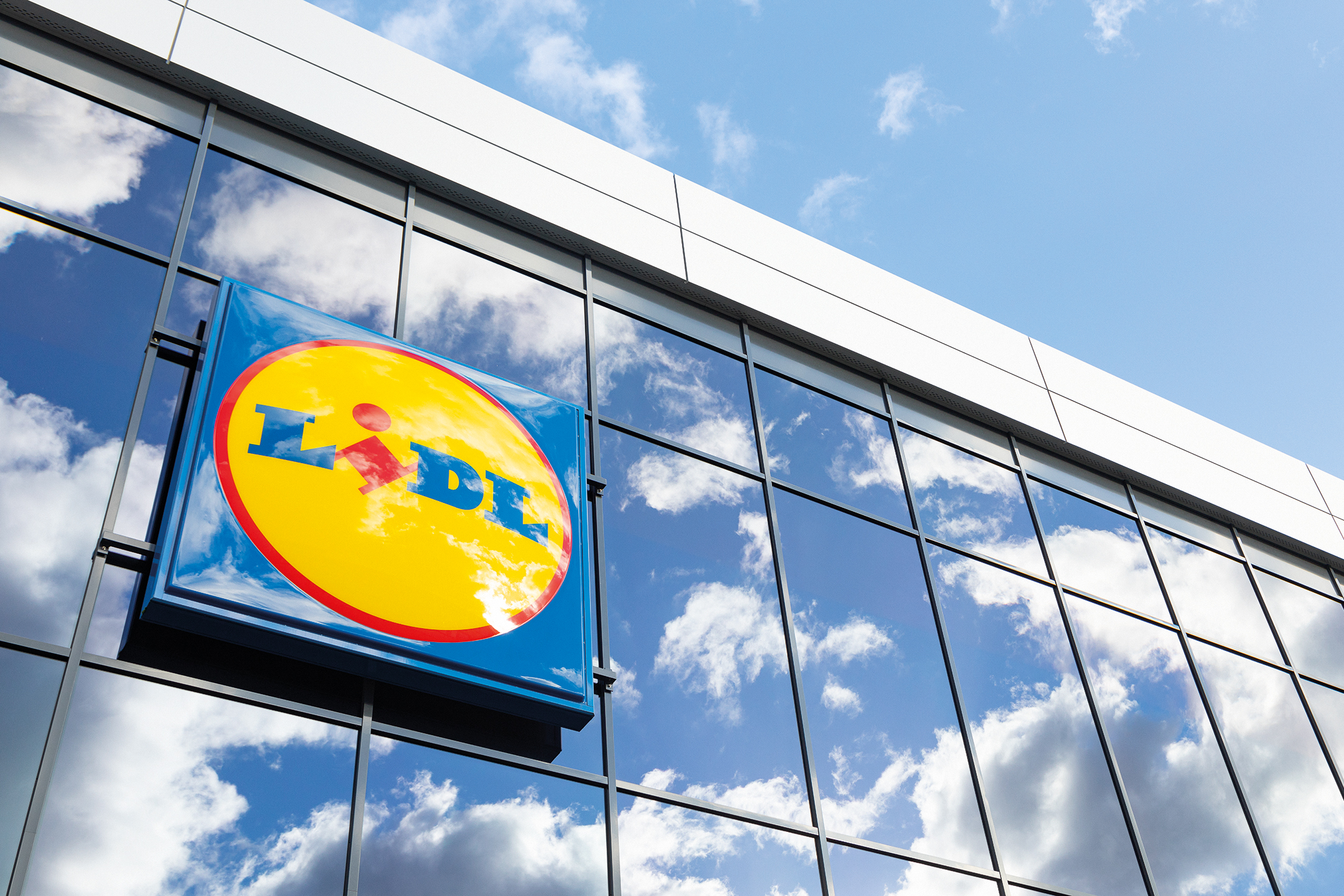 Надпис Lidl на стъклена фасада на сграда, с отражение на небе и облаци.
