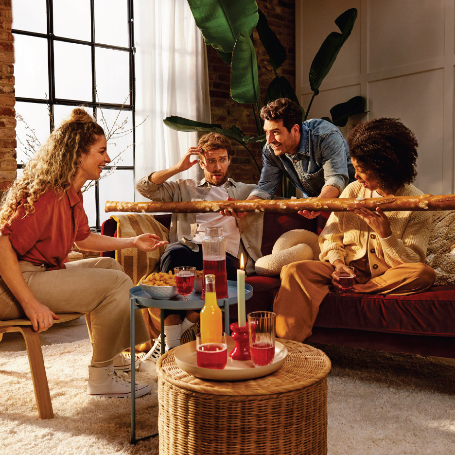 Четирима приятели споделят дълъг солен геврек и напитки на диван в уютна всекидневна.