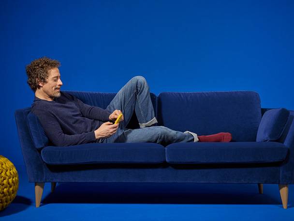 Мъж се отпуска на син диван с мобилен телефон.