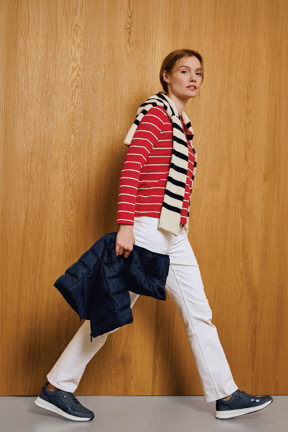 Жена в червена раирана блуза, бели панталони и тъмносини маратонки, държи тъмносиньо яке.