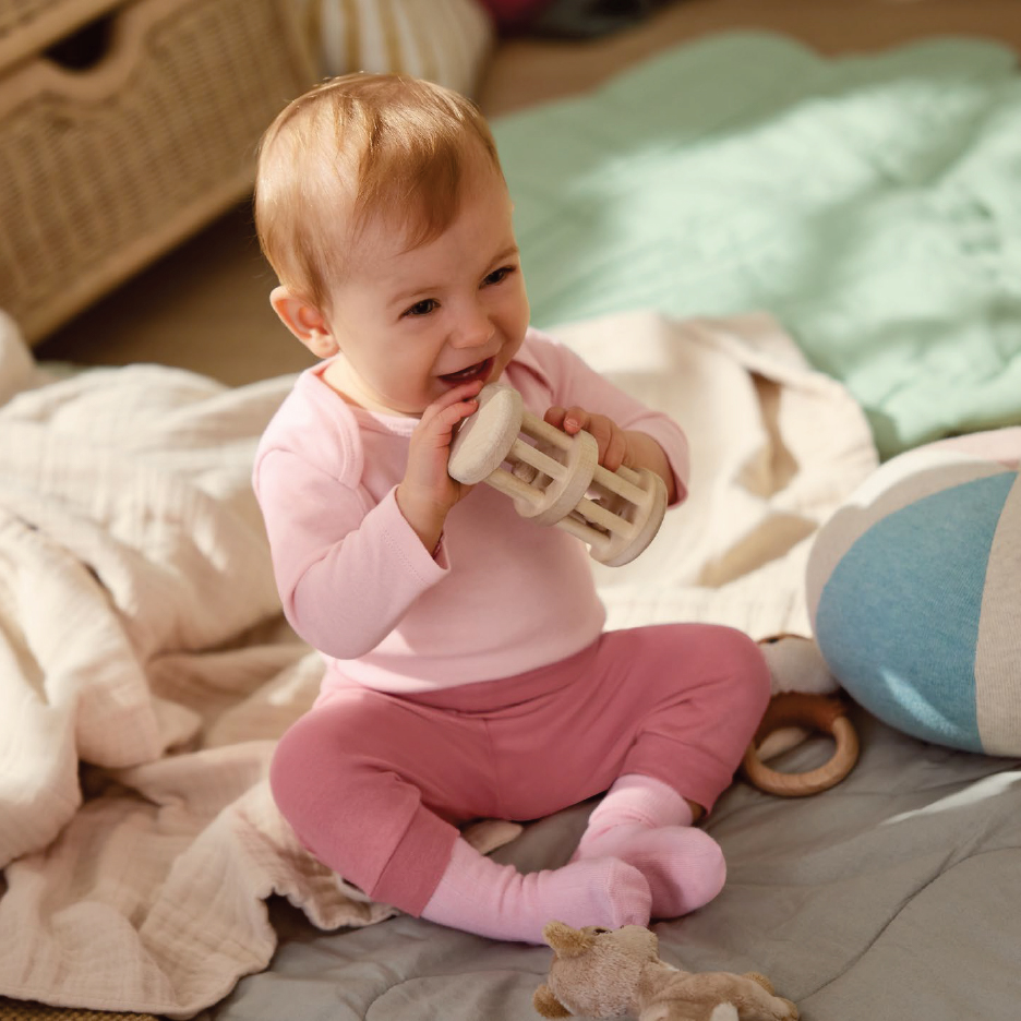 Усмихнато бебе в розови дрехи, седнало на одеяло, играе с дървена дрънкалка.