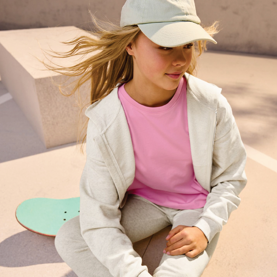 Момиче с розова тениска, сив суичър и шапка, седнало до скейтборд.