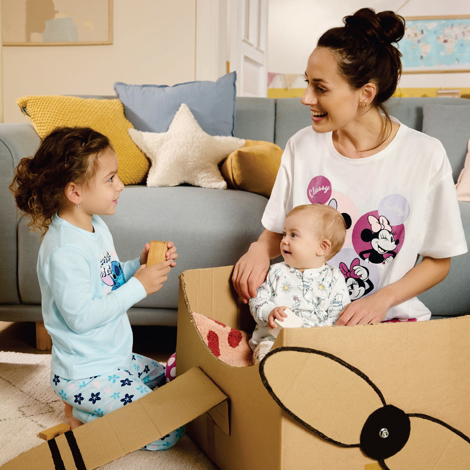 Майка и две деца си играят с картонен самолет, облечени в пижами.