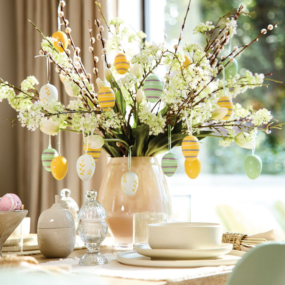 Великденска украса за маса с ваза, върбови клонки, цветя и висящи великденски яйца.
