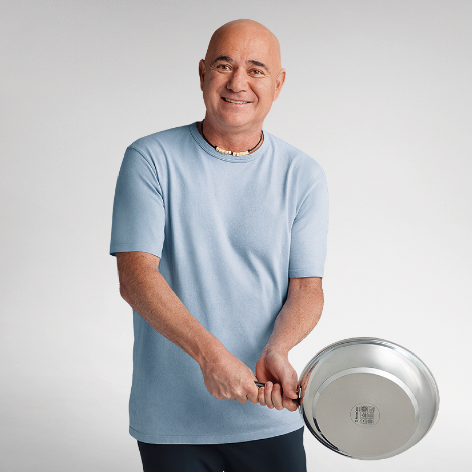 Андре Агаси в синя тениска държи тиган, усмихвайки се на камерата.