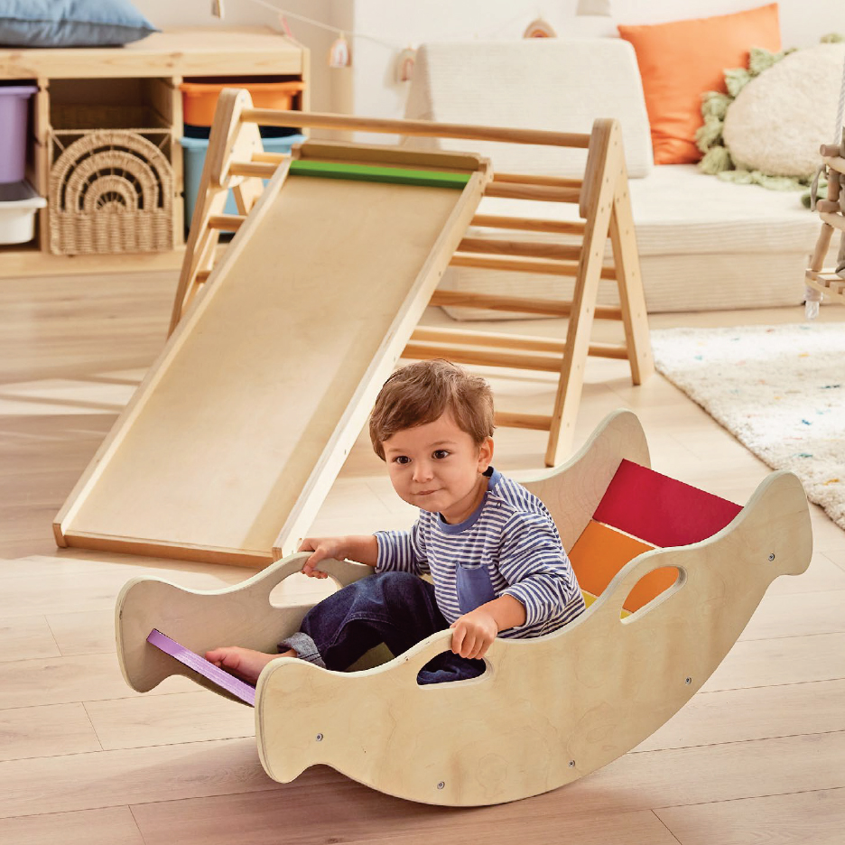 Малко дете седи в дървена люлка, докато зад него има дървена пързалка и катерушка.