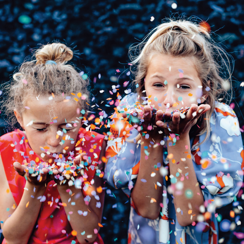 Две момичета духат конфети от ръцете си, усмихнати и щастливи.