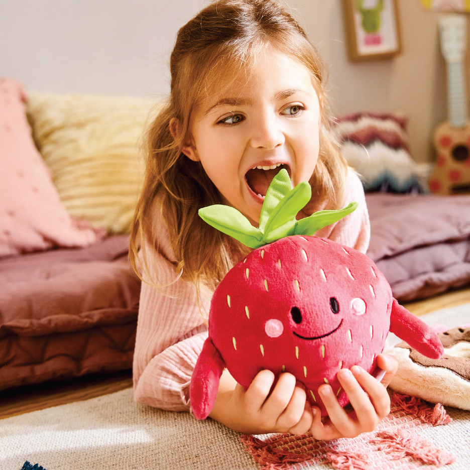 Момиче си играе с плюшена ягода, докато лежи на пода.