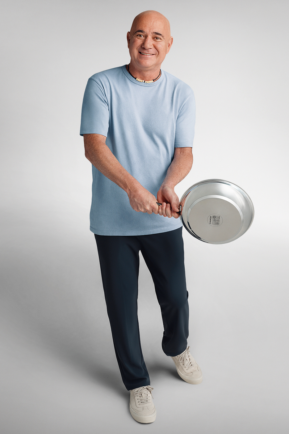 Мъж в синя тениска и тъмни панталони държи тиган, усмихвайки се.