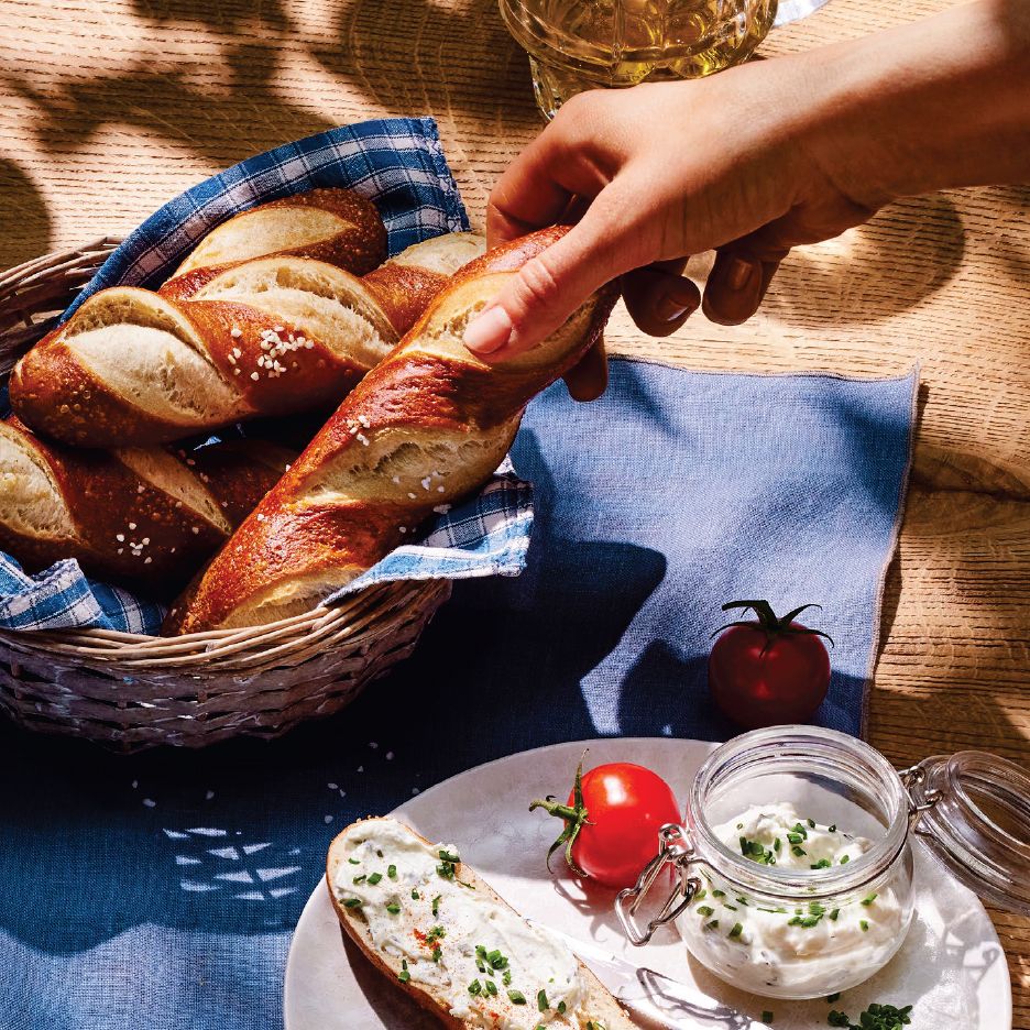 Ръка посяга към кошница с пресни солени пръчици, докато на масата има домат и буркан със сирене.