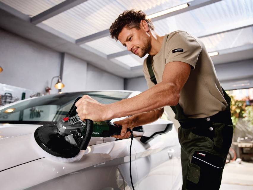 Мъж полира бял автомобил с електрическа полираща машина в гараж.