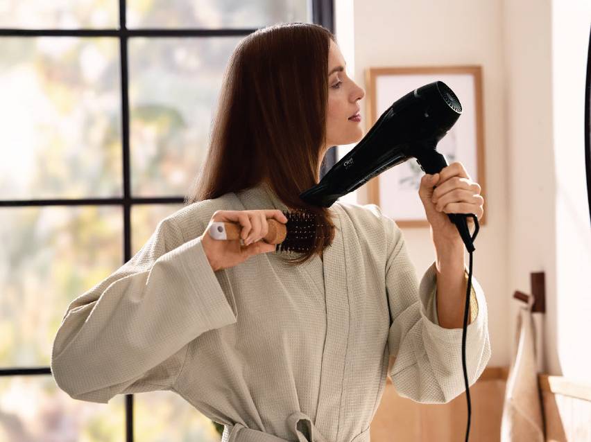 Жена суши косата си със сешоар и четка за коса в банята.