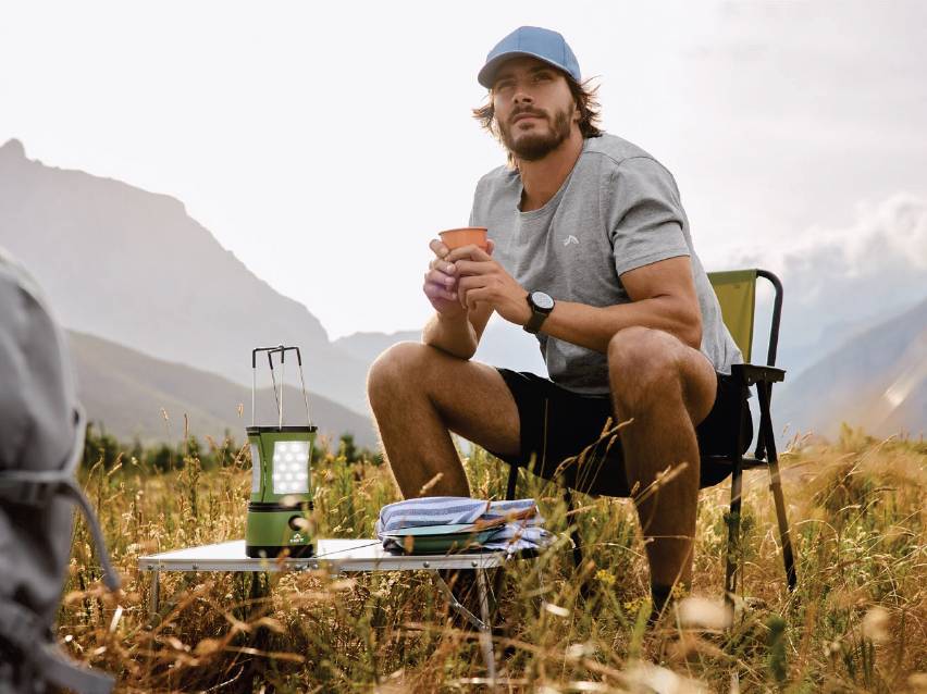 Мъж на къмпинг стол с чаша, къмпинг фенер и маса сред природата.