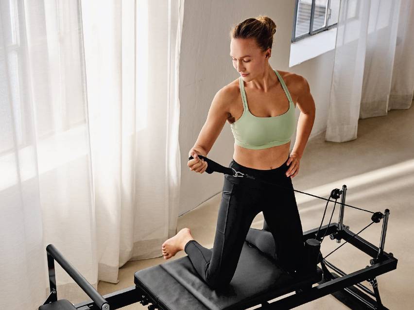 Жена тренира пилатес на реформатор, облечена в спортен сутиен и клин.