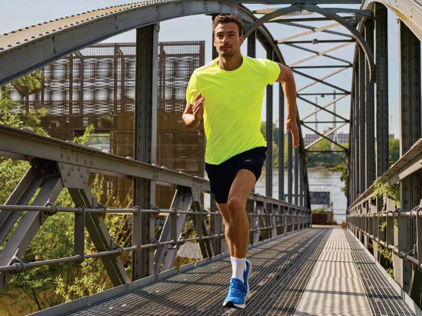 Мъж в неоновожълта тениска и тъмносини шорти тича по метален мост.
