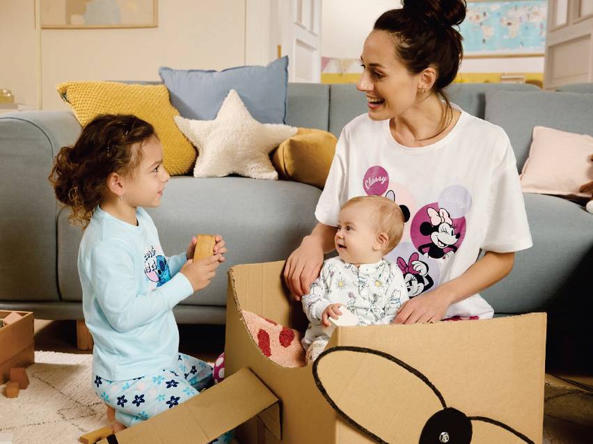 Майка и две деца в пижами си играят с картонена кутия в хола.