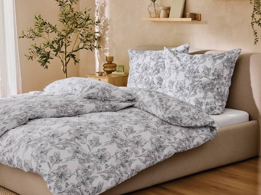 Спалня с бял спален комплект с флорални мотиви, нощно шкафче и растение.