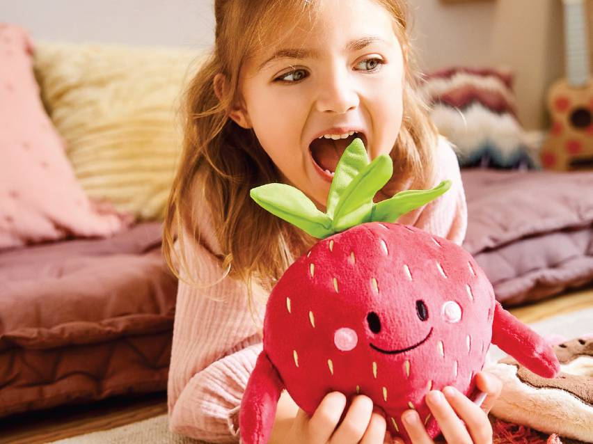 Момиче си играе с плюшена ягода, отваряйки уста, сякаш ще я изяде.
