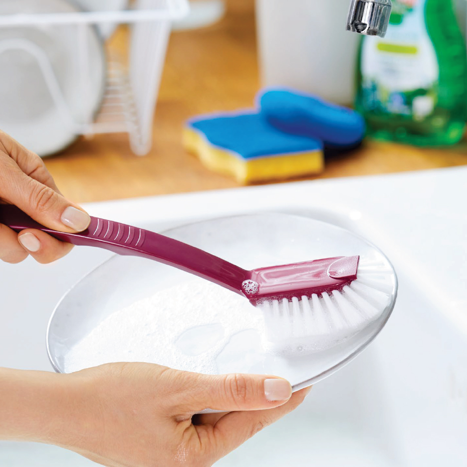Ръце мият чиния с четка за съдове, на заден план гъби и препарат за миене.