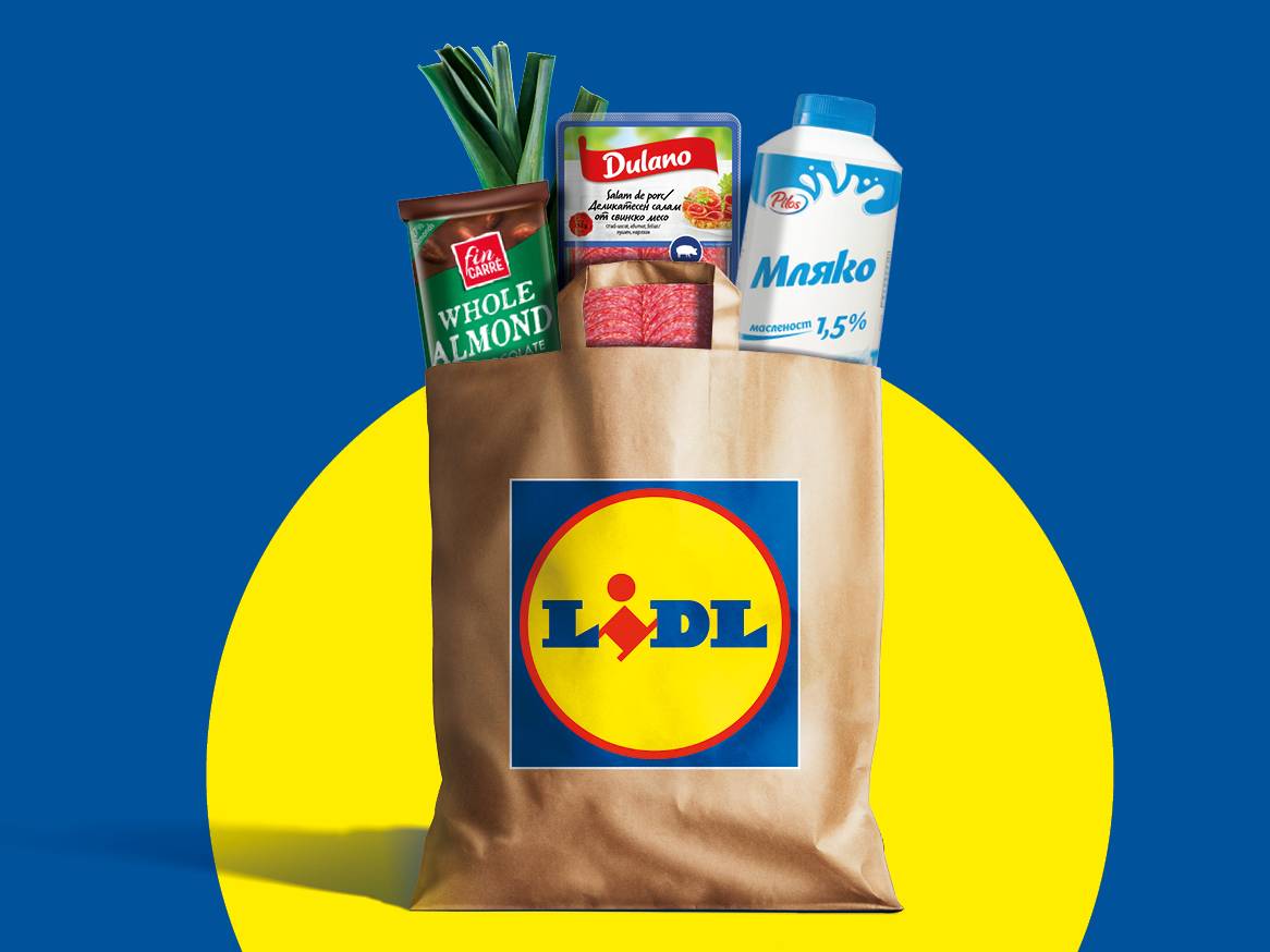 Хартиена торба с хранителни стоки, включваща мляко, салам, шоколад с бадеми и праз, на син фон с жълт кръг.