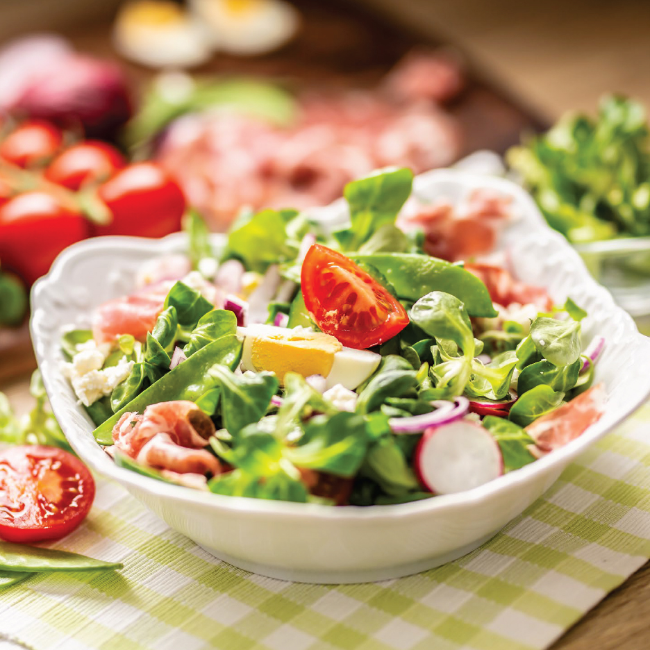 Салата от маруля с домати, яйца, шунка, червен лук и репички в бяла купа.