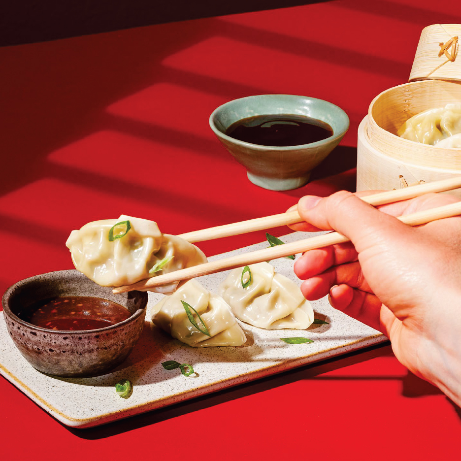 Ръка с клечки държи кнедли над сос, с още кнедли и бамбуков съд на червена маса.